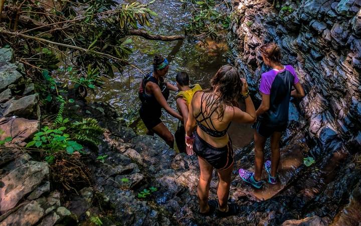 A group of young people wearing swimwear descend a rocky incline toward a body of water. 
